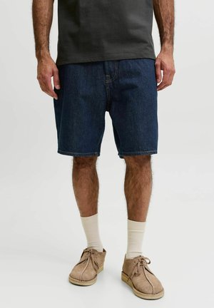 Homme portant un short en jean bleu foncé, des chaussures en daim beige avec lacets, des chaussettes blanches et un t-shirt noir, debout sur fond blanc.