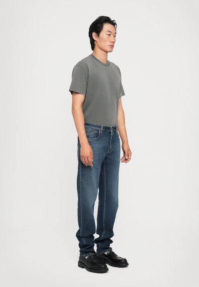 Un tricou din bumbac gri asortat cu blugi din denim albastri. Modelul poartă mocasini negri. Design simplu și minimalist, cu croială dreaptă.