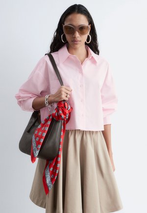 Vrouw draagt een oversized roze blouse met knopen en een beige rok, grote creolen, zonnebril, draagt een donkerbruine tas met een rood patroon sjaal.