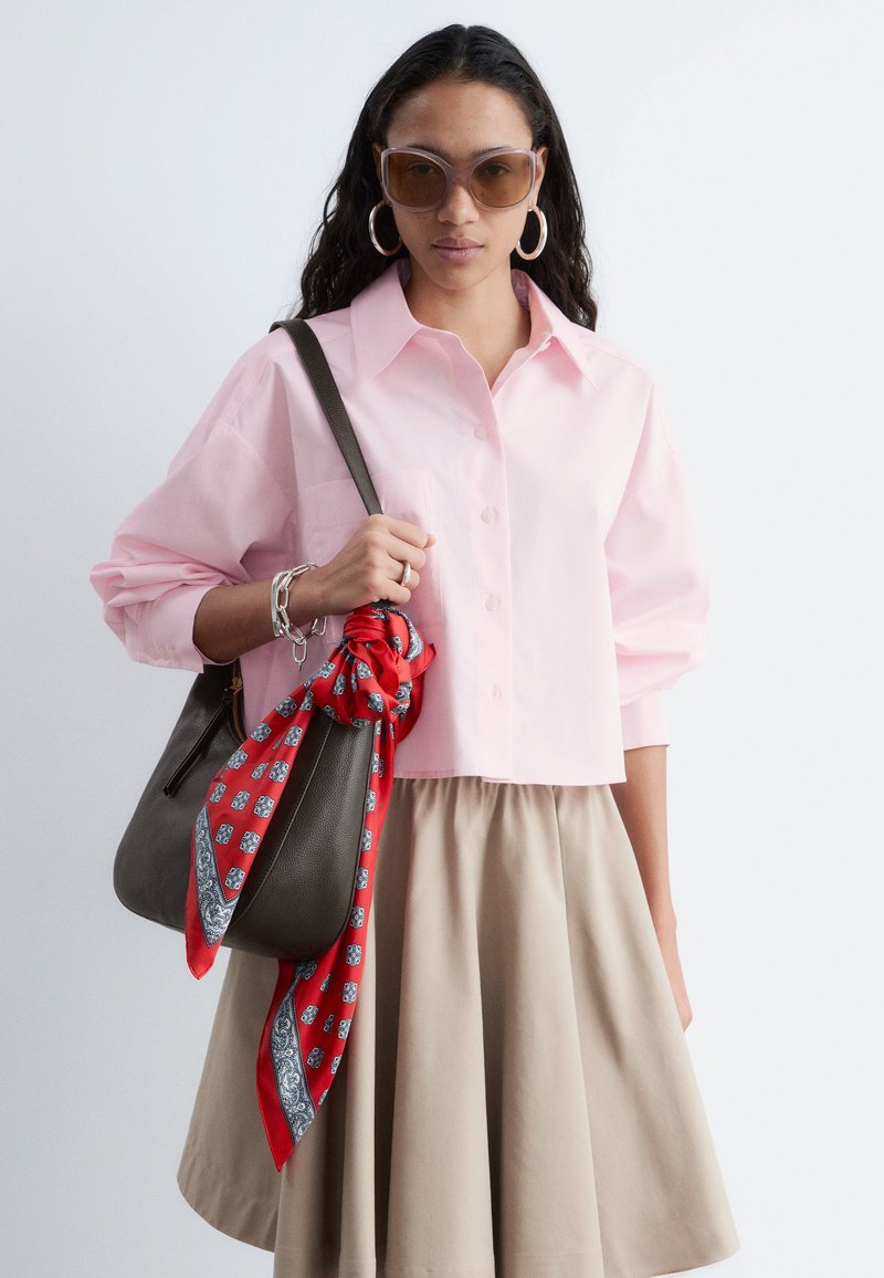 Mujer con camisa rosa de botones de gran tamaño y falda beige, pendientes grandes de aro, gafas de sol, llevando un bolso marrón oscuro con pañuelo rojo con estampado.