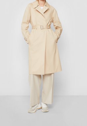 Mujer con gabardina beige con cinturón, pantalones de pierna ancha y zapatillas blancas, de pie frente a un fondo gris liso.