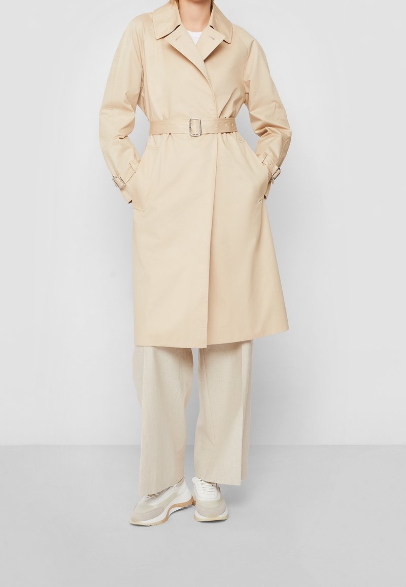Femme portant un trench-coat beige ceinturé avec un pantalon à jambes larges et des baskets blanches, debout devant un fond gris uni.