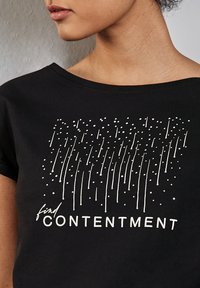 Žena v černém tričku s bílými vertikálními pruhy a tečkami nad nápisem "najdi SPOKOJENOST" na přední straně.