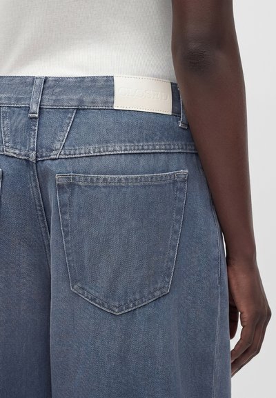 Vue arrière d’une personne portant un jean en denim bleu clair avec une étiquette blanche « CLOSED » à la taille et une grande poche arrière.