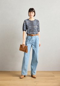 Kortermet, stripete topp i svart og blått, høytlivs lyseblå jeans, brun belte, holder en sandfarget semsket clutch, beige sko.