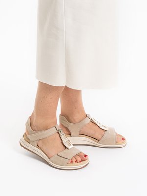 Sandalias de ante beige con punta abierta, correas ajustables y adornos metálicos decorativos, combinadas con pantalones blancos de pierna ancha.