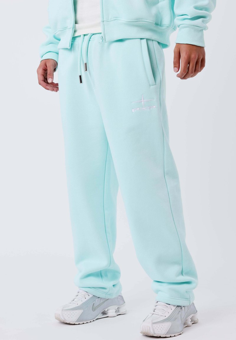 Uomo che indossa pantaloni tuta azzurri chiari con logo PXP e sneaker bianche-grigie Nike, in piedi contro uno sfondo chiaro e semplice.