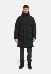 Parka negra con capucha, múltiples bolsillos frontales y una textura mate elegante. Combinada con jeans oscuros y botas negras.