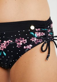 Schwarze Bikinischlüpfer mit weißen Punkten und einem floralen Muster in Pink und Grün. Mit seitlicher Raffung und Kordelzugverschluss sowie einem Logo-Detail.