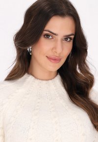 Pullover bianco lavorato a maglia con un motivo a treccia testurizzato, accentuato da piccole perle. Il modello ha capelli lunghi e ondulati e indossa orecchini d'argento.