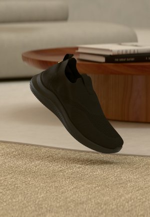 Μαύρο αθλητικό παπούτσι χωρίς κορδόνια που αιωρείται πάνω από ένα μπεζ υφαντό χαλί, με ένα ξύλινο τραπεζάκι σαλονιού και βιβλία στο φόντο.