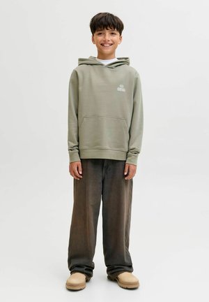 Ragazzo in piedi e sorridente, indossa un felpa verde chiaro, pantaloni larghi scuri e scarpe beige su uno sfondo bianco semplice.