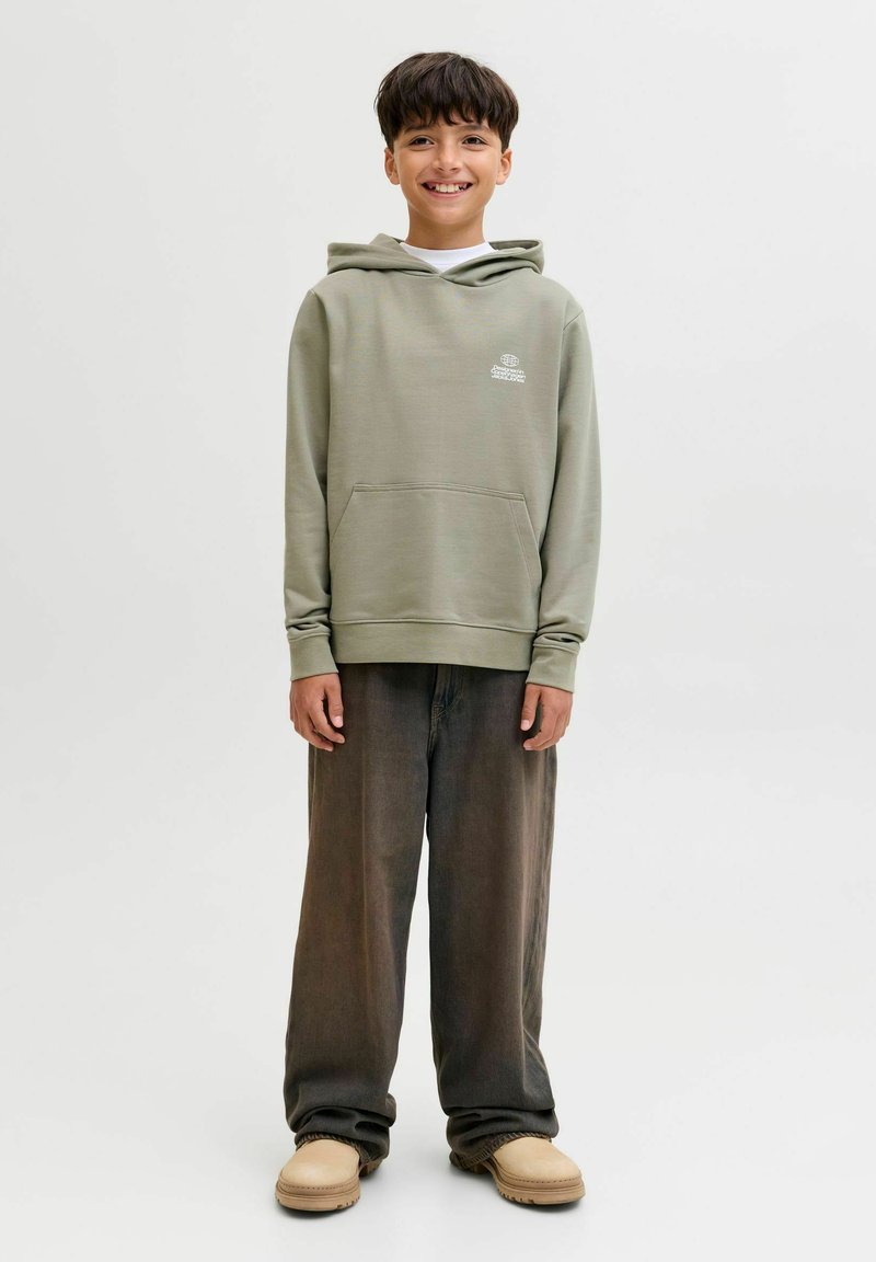 Chlapec stojí a usmieva sa, má na sebe svetlozelenú mikinu, tmavé voľné nohavice a béžové topánky na pozadí čistej bielej steny.