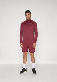 Bordeauxrotes Langarm-Athletic-Oberteil mit einem halben Reißverschluss, kombiniert mit passenden Shorts. Mit einem Logo auf der Brust und in Kombination mit schwarzen Sneakers.