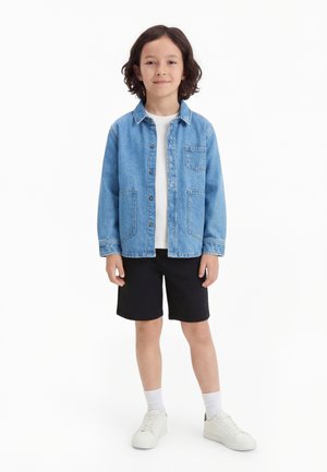 Dieťa s tmavými vlasmi po plecia stojí v modrej džínsovej bunde, bielom tričku, čiernych šortkách, bielych ponožkách a bielych teniskách.