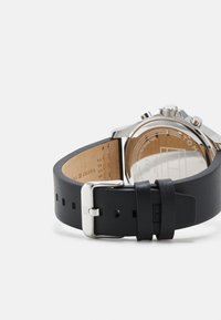 Correa de reloj de cuero negro con hebilla de metal plateado, mostrando un interior de color marrón claro. Caja de acero inoxidable con respaldo transparente y características de cronógrafo.