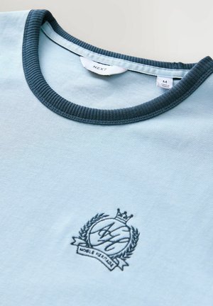 Svetlomodré tričko vyrobené z bavlny. Má tmavomodrý rebrovaný golier a vyšívanú emblému s korunou a vavrínovými detailmi.