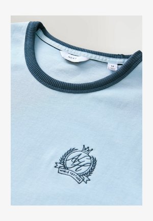 Hellblaues T-Shirt aus Baumwolle. Verfügt über einen dunkelblauen Rippkragen und ein gesticktes Emblem mit Krone und Lorbeerdetails.