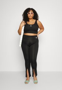 Crop top sportivo nero e leggings a vita alta con fessure laterali. Il modello indossa sandali aperti verde menta e tiene una cintura bianca con buchi.