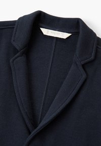 Tmavě modrý blejzr z texturované látky, vybavený klopou s výřezem a zdobenými stehy podél límce a přední části. Viditelná značka značky.