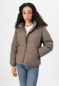 Giacca a vento marrone con zip frontale, colletto alto e polsini elasticizzati. Il design trapuntato presenta cuciture orizzontali e tasche laterali.