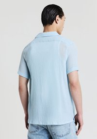 Uomo con capelli scuri slicked-back che indossa una camicia a maniche corte azzurro chiaro a trama e jeans in denim chiari, di spalle su sfondo neutro.
