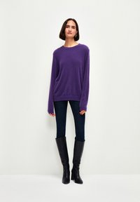 Lila, weicher Strickpullover mit lässiger Passform und halblangen Ärmeln, kombiniert mit blauen engen Jeans und knielangen schwarzen Stiefeln.
