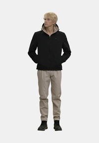 Schwarze Jacke mit Reißverschluss und hohem Kragen, beigem Futter und grünem Logo, kombiniert mit hellbeigen Hosen und schwarzen Knöchelstiefeln.