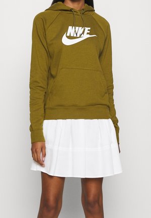 Osoba w oliwkowozielonej bluzie z kapturem Nike z białym logo i białej plisowanej spódnicy, stojąca na neutralnym jasnym tle.