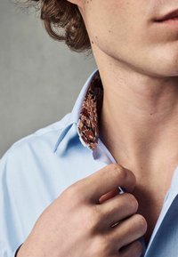 Cămașă bleu deschis cu guler căptușit cu un model floral. O mână își ajustează gulerul, care are detalii de țesătură contrastante.