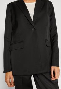 Black blazer made of smooth fabric, featuring a single button, two front pockets, and structured lapels. Tailored fit with long sleeves.