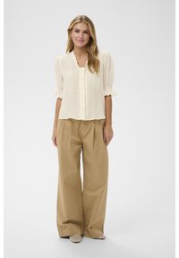 Mlada ženska, oblečena v kremasto bluzo z balonastimi rokavi in širokimi hlačami kamelj barve, stoji pred enobarvnim svetlim ozadjem.