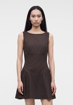 Jeune femme aux longs cheveux noirs portant une robe moulante sans manches marron foncé, debout devant un fond clair uni.