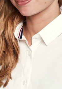 Biała koszula na guziki z kontrastującym kołnierzem w paski w kolorze granatowym i czerwonym. Miękka tkanina o gładkiej fakturze, widok z bliska na dekolt.