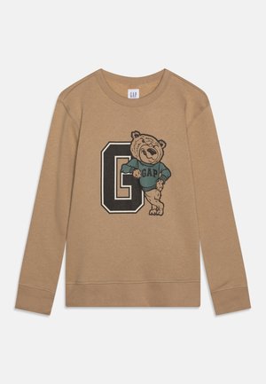Hellbraunes Langarm-Sweatshirt mit einem großen schwarzen "G" und einem lächelnden Bären, der einen blauen GAP-Pullover trägt und sich an den Buchstaben lehnt.