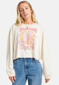 Cream-colored cropped long-sleeve t-shirt featuring a colorful graphic print of a bird and flowers. Paired with high-waisted blue jeans.