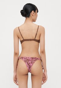 Rudas maudymosi liemenėlė su reguliuojamomis petnešėlėmis, derinama su rožinėmis bikini kelnaitėmis su gėlėmis, šoninėmis raišteliais ir žemu kirpimu.