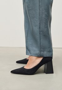 Crne plitke cipele od antilopa s visokom petom, šiljastim vrhom i masivnom crnom petom. Uparene s svijetloplavim, skraćenim hlačama širokih nogavica.