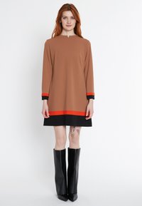 Braunes Langarmkleid mit einem leicht hohen Kragen, rot-schwarz gestreiftem Saum und Manschetten, kombiniert mit glänzenden schwarzen knielangen Stiefeln.