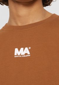 Camiseta de algodón marrón con un logo impreso en blanco, "MA MARTIN ASBJØRN," situado en la parte izquierda del pecho. Diseño de cuello redondo.