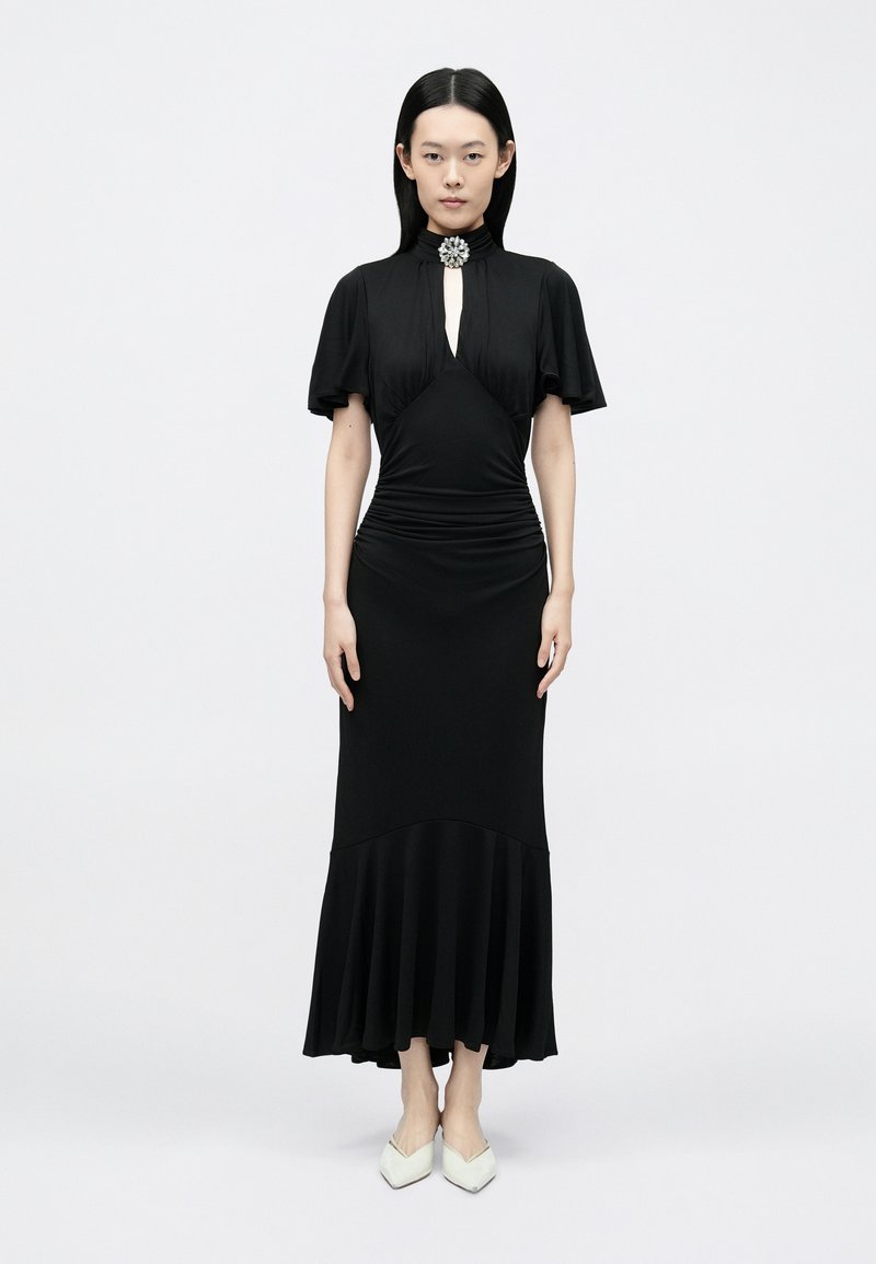 Fekete ruha rövid, fodros ujjakkal, kulcslyuk nyakkivágással, deréknál ráncolt, és bővülő szegéllyel. Világos színű, hegyes orr�ú cipővel párosítva.