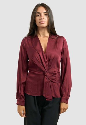 Blusa de satén burdeos con manga larga, escote en V profundo y detalle de nudo en la parte frontal. Textura suave y ligero brillo.