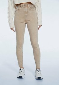 Pantalones vaqueros ajustados de talle alto en color beige, con una textura suave y un ligero estiramiento, que cuentan con un cierre de botón en la parte delantera y aberturas en los tobillos. Combinados con zapatillas blancas.