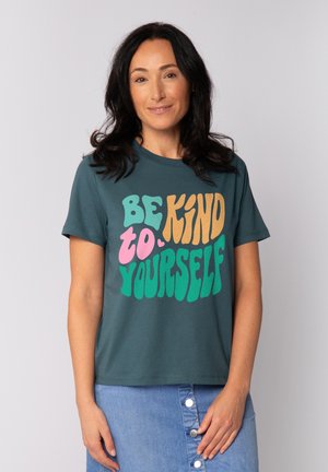 Frau mit dunklem Haar, die ein türkisfarbenes T-Shirt mit buntem Text "Sei freundlich zu dir selbst" und einen blauen Rock mit Knöpfen trägt, steht vor einem schlichten Hintergrund.