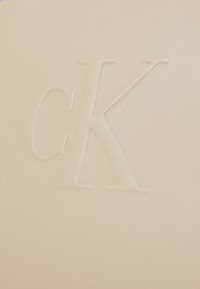 Beżowy skórzany portfel z tłoczonym logo "CK", gładką fakturą i subtelnym połyskiem na powierzchni. Bez dodatkowych ozdób.