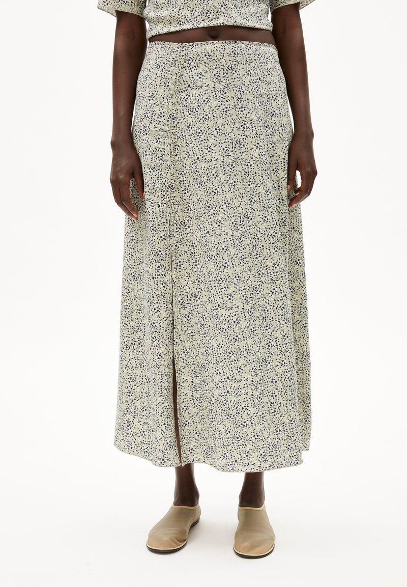Maxi-jurk van een lichtgroene stof met een verspreide marineblauwe print, heeft een voorkant-split en een elastische tailleband. Gecombineerd met beige instapschoenen.