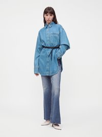 Femme portant une chemise en denim bleu oversize avec une ceinture noire, un jean bleu délavé et des talons pointus blancs, debout devant un fond blanc uni.