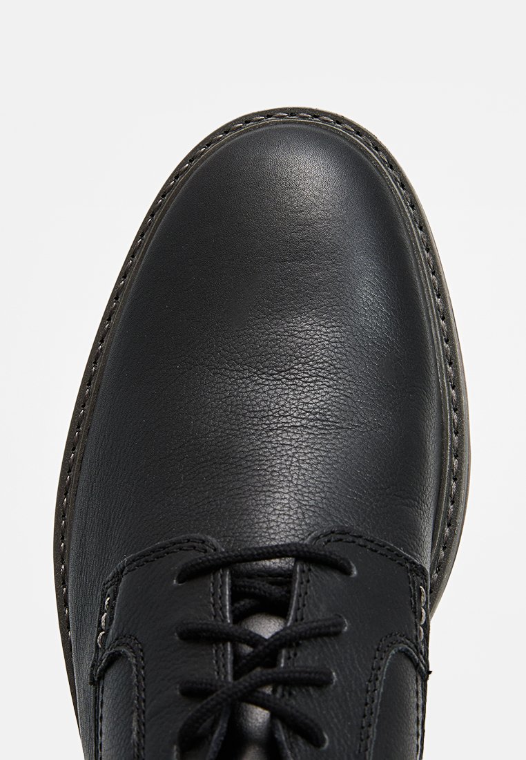 Scarpa in pelle nera con una texture liscia, punta arrotondata e dettagli cuciti. Presenta lacci e una suola robusta per il supporto.