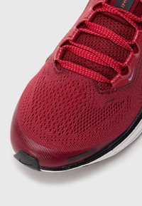 Chaussure de sport rouge avec un dessus en maille, dotée d'un tissu texturé, de six lacets rouges et d'un capuchon en caoutchouc noir avec une semelle blanche.