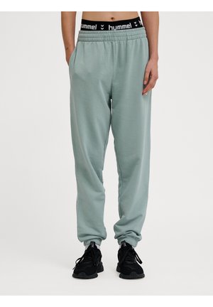 Person iført lysegrønne joggebukser med strammet ankler, sort linning med "hummel" logo, sorte sneakers og hænderne i lommerne.